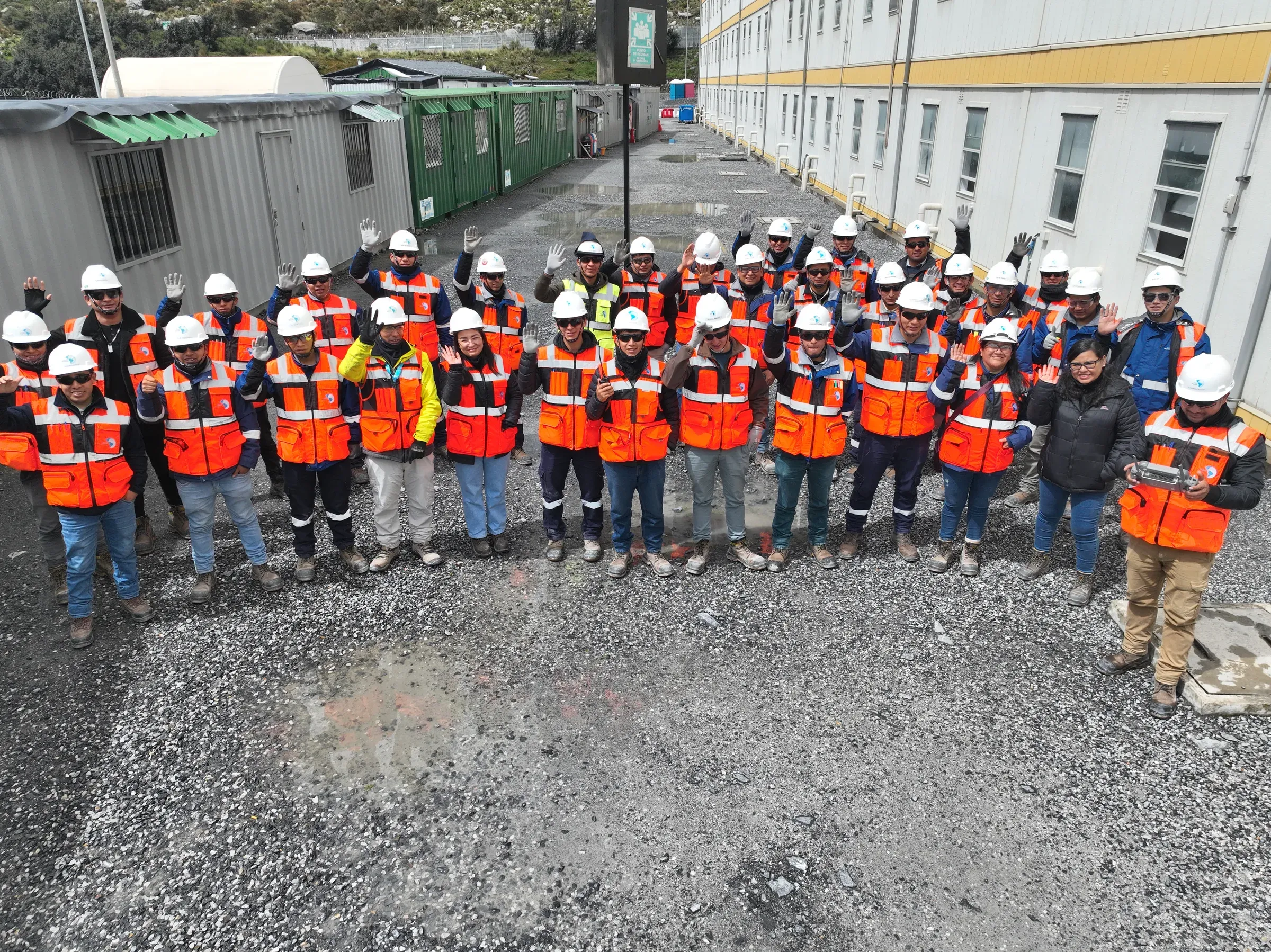 Grupo numeroso de personal de Global Mapping con chalecos naranja y cascos blancos, saludando entre los módulos de un campamento